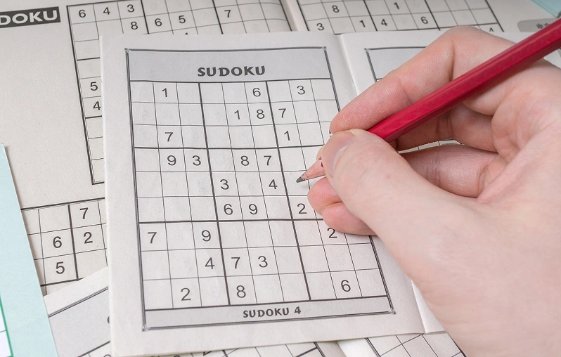 Nahaufnahme einer rechten Hand, die einen roten Bleistift hält und eine Zahl in ein Sudoku-Gitter einträgt. Im Hintergrund liegen weitere aufgeschlagene Rätselseiten.