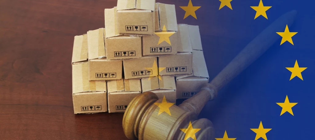 Eine aufgestapelte Pyramide aus kleinen braunen Pappkartons auf einem Holztisch. Im Vordergrund liegt ein Richterhammer aus Holz, während die rechte Bildhälfte von der blauen Flagge der Europäischen Union mit ihren gelben Sternen überlagert wird – symbolisch für das neue Urteil von der EU zum Wegfall der Zollbefreiung für Kleinsendungen