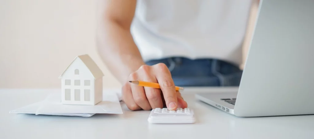 Steuerberater berechnet Vorsteueraufteilung bei gemischt genutztem Gebäude – Miniaturhaus, Taschenrechner und Laptop auf Schreibtisch